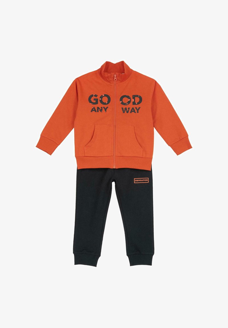 Veste zippée orange avec le texte "GO OD ANY WAY", associée à un pantalon de survêtement noir avec un petit patch logo orange. Tissu doux, design décontracté.