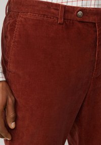 Roestkleurige corduroy broek met verticale ribbelstructuur, voorzien van een bijpassende tailleband en een metalen knoopsluiting. Hand zichtbaar aan de zijkant.
