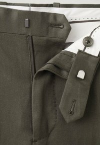 Zbliżenie na pas spodni w oliwkowym kolorze, pokazujący metalowy haczyk, guzik, zamek błyskawiczny oraz wewnętrzną białą podszewkę z czarnymi detalami szwów.