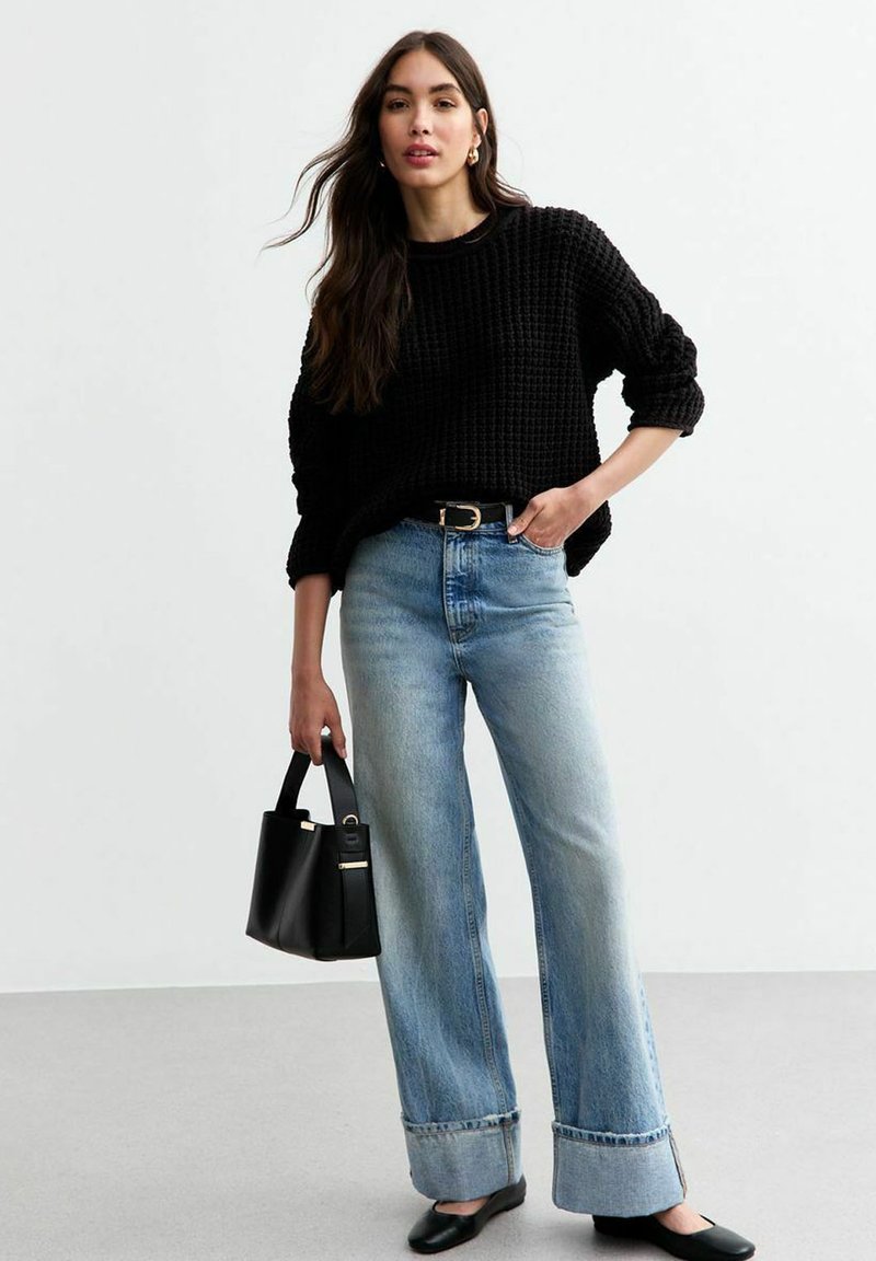 Zwarte gebreide trui, lichtblauwe wijde jeans, zwarte flats en een kleine zwarte handtas met gouden hardware. Gevarieerde materialen en een losse pasvorm.