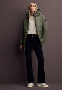 Doudoune vert olive avec capuche, associée à un haut beige clair, un jean foncé et des baskets blanches. La doudoune est dotée d'une fermeture zippée et de poches latérales.