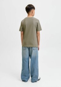 Dječak stoji okrenut leđima, obučen u maslinasto zeleni majicu, široke svijetloplave traperice i bijele tenisice, nasuprot jednobojnoj pozadini.