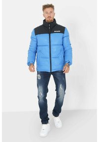 Blauwe pufferjack met zwarte kraag en rits, met een glanzende afwerking. Gekocht met versleten donkere jeans en witte sneakers.