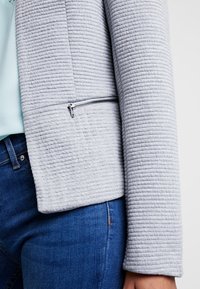 Nahaufnahme einer Person, die eine hellgraue strukturierte Jacke mit einer horizontalen Reißverschlusstasche und blauen Jeans trägt, vor einem schlichten Hintergrund.