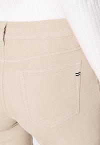 Pantaloni in cotone beige misto con una tasca posteriore che presenta una sottile striscia nera orizzontale. Il tessuto ha un aspetto liscio e strutturato.