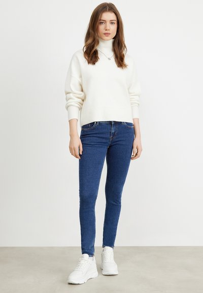 Suéter de cuello alto color crema, ajuste relajado con hombros caídos, combinado con jeans ajustados azul oscuro y zapatillas blancas.