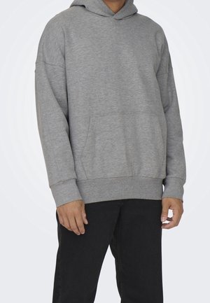 Personne portant un sweat à capuche gris ample avec une poche avant et un pantalon noir, présenté de la nuque jusqu'au milieu des cuisses sur un fond blanc.
