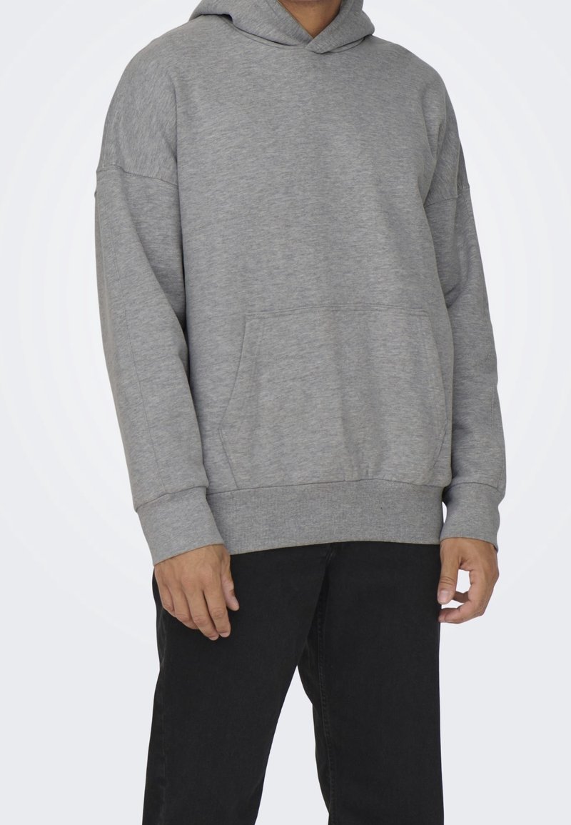 Personne portant un sweat à capuche gris ample avec une poche avant et un pantalon noir, présenté de la nuque jusqu'au milieu des cuisses sur un fond blanc.