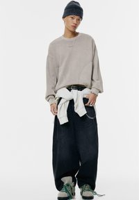 Felpa oversized beige con spalle scese, jeans neri larghi, un hoodie bianco legato in vita e sneakers grigie con lacci verdi.