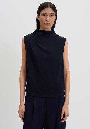 Femme aux longs cheveux foncés portant un haut noir sans manches à col haut et un pantalon noir, debout devant un fond uni clair.
