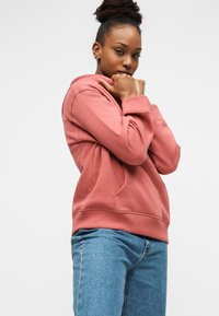 Ροζ φούτερ με απαλό ύφασμα, τσέπη καγκουρό, ριμπ μανσέτες και μέση, σε συνδυασμό με τζιν παντελόνι.