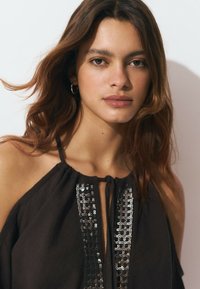 Haut sans manches noir avec un col froncé et un accent métallique décoratif sur le devant. Tissu doux, texture fluide et liens au cou.