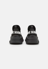 Zapatillas deportivas negras con parte superior de malla, suela gruesa y acolchada, y grandes letras blancas en el talón. La vista trasera muestra una lengüeta y textura.