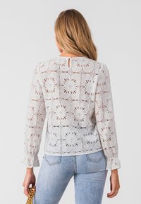 White lace blouse with floral pattern, long sleeves, ruffled cuffs, keyhole back detail, paired with light blue jeans.