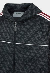 Giacca nera con zip, caratterizzata da un motivo testurizzato, una striscia bianca solida e accenti rossi sulle spalle. Il materiale sembra liscio e traspirante.