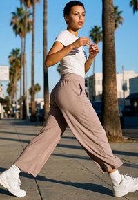 Young person walking briskly on sunlit urban sidewalk lined with tall palm trees, wearing white shirt, beige pants, and white sneakers.