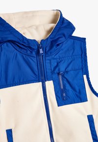 Blauwe en crèmekleurige mouwloze vest met capuchon, voorzien van doorgestikt materiaal, een ritssluiting aan de voorkant en contrasterende kleurdetaillering in de zakken.