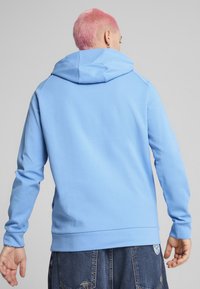 Sudadera azul clara hecha de un tejido suave, con capucha y mangas largas. La vista trasera muestra un diseño sin costuras y un ajuste ceñido en la cintura.