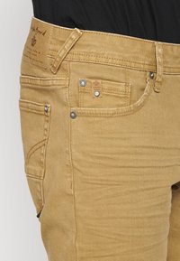 Kaki denim teksad, millel on reljeefne viimistlus, viis taskut, metallaktsendid ja kaubamärgiga nahkpaan vöökoosal.