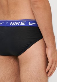Schwarze Schwimmbriefs für Männer mit einem blauen elastischen Bund, der mit weißem "NIKE"-Branding versehen ist. Glatte Textur, enganliegendes Design.