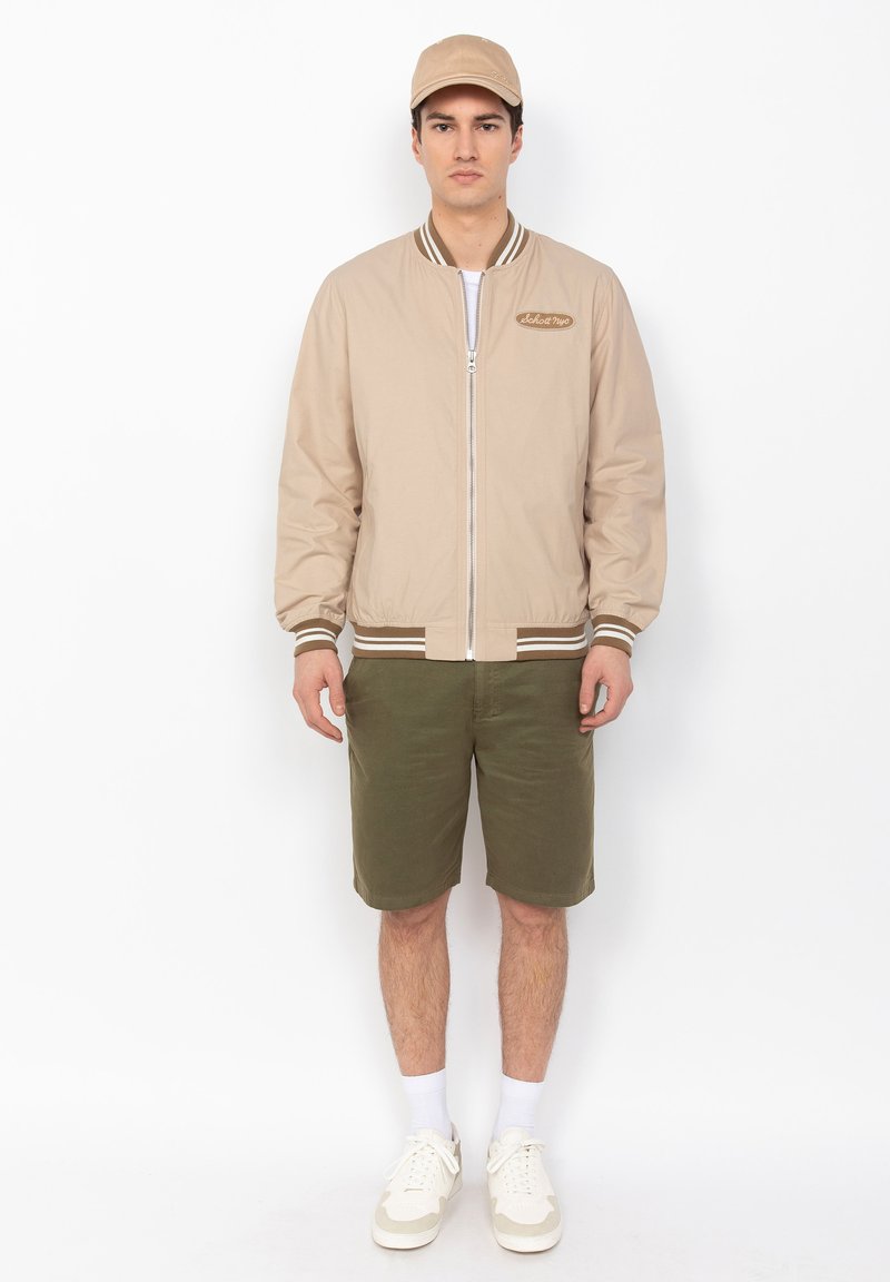 Beige Bomberjacke mit Reißverschluss, geripptem Kragen und braunen Streifen an den Bündchen, kombiniert mit olivgrünen Shorts und weißen Sneakers.