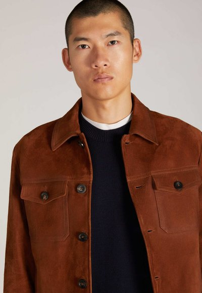 Braune Wildlederjacke mit Kragen, zwei Brusttaschen und schwarzen Knopfakzenten. Über einem marineblauen Strickpullover getragen. Glatte Textur, taillierte Passform.