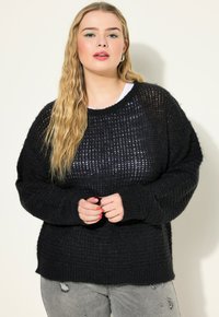 Schwarzer gestrickter Pullover mit lockerer, strukturierter Webart und gerippten Bündchen, getragen über einer weißen Schicht. Kombination mit abgewetzten grauen Jeans.