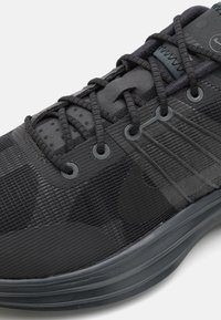 Gros plan d'une chaussure de sport noire avec tissu texturé, lacets intégrés et couture visible de la semelle sur fond blanc.
