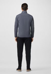 Hombre de pie de espaldas, vistiendo una camiseta de manga larga gris, pantalones negros y zapatillas negras, frente a un fondo claro y liso.