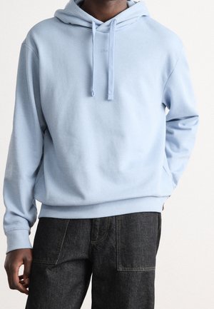 Man wearing light blue hoodie with drawstrings and black jeans, standing against a plain white background.