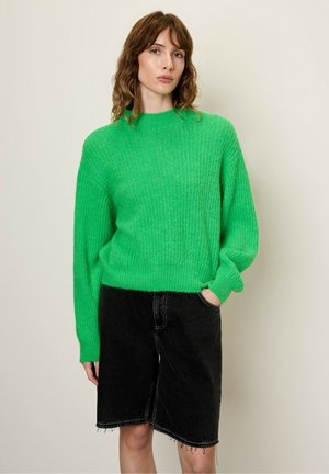 Grüner Strickpullover mit hohem Kragen und gerippter Textur, kombiniert mit schwarzen Jeansshorts mit ausgefransten Kanten und seitlichen Taschen.