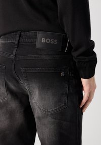 Jeansi negri din denim au o textură netedă, croi drept și o etichetă din piele cu inscripția „BOSS” pe talie. Detalii subtile de decolorare sunt vizibile.