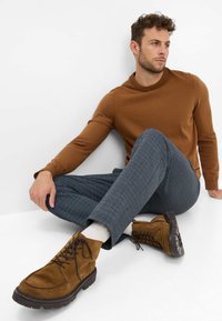 Maglione marrone a maglia, pantaloni a quadretti blu e stivali in camoscio marrone con suole in gomma nera, seduto su uno sfondo bianco.