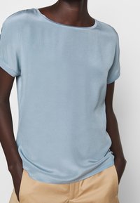 Personne portant un chemisier en satin bleu clair à manches courtes avec un col rond et un pantalon beige, les mains dans les poches, sur un fond uni.