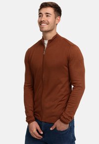 Brun zip-up genser med teksturert strikk, høy krage og lange ermer, kombinert med blå jeans. Har en glidelås som hoveddetalj.