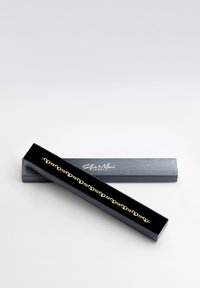 Armband i guld med hjärtlänk som visas i en svart smyckeslåda med en silverlogo. Lådan har en texturerad yta som framhäver armbandet.