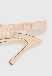 Sandale à talons hauts beige avec une finition brillante, ornée de strass sur le côté et d'un détail en nœud en tissu sur la bride de la cheville.