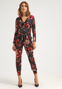 Jumpsuit nera floreale con maniche lunghe, scollo a corona e vita con cintura. Presenta grandi fiori rossi e rosa su una finitura opaca.