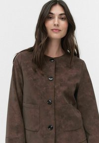 Jeune femme aux longs cheveux bruns portant une veste en daim marron avec des boutons et deux poches devant, se tenant contre un fond clair.
