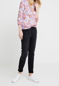 Staande vrouw in een paarse bloemenblouse, zwarte opgerolde jeans, witte sneakers en gouden ringen om haar linkerhand.