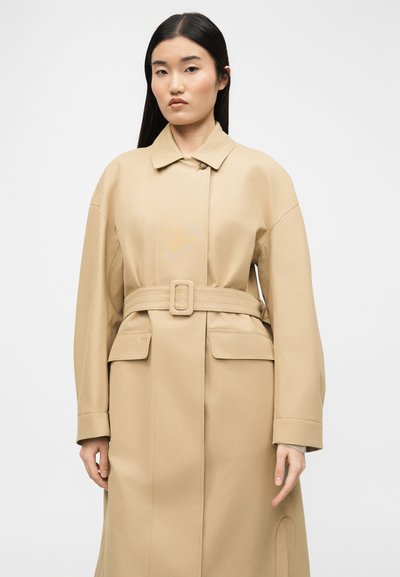 Γυναίκα με μακριά ίσια μαλλιά που φοράει μια μπεζ καμπαρντίνα με ζώνη και τσέπες με καπάκι, που στέκεται μπροστά σε ένα απλό φωτεινό φόντο.