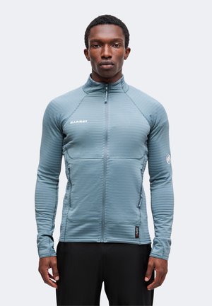 Mann trägt eine hellblaue Jacke mit durchgehendem Reißverschluss und zwei vorderen Reißverschlusstaschen sowie schwarze Hose, blickt neutral nach vorne.