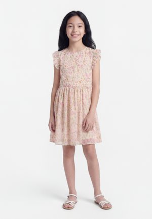 Κορίτσι που στέκεται και χαμογελά, φοράει ανοιχτό ροζ φόρεμα με λουλούδια και μανίκια με βολάν και άσπρα σανδάλια, σε απλό λευκό φόντο.