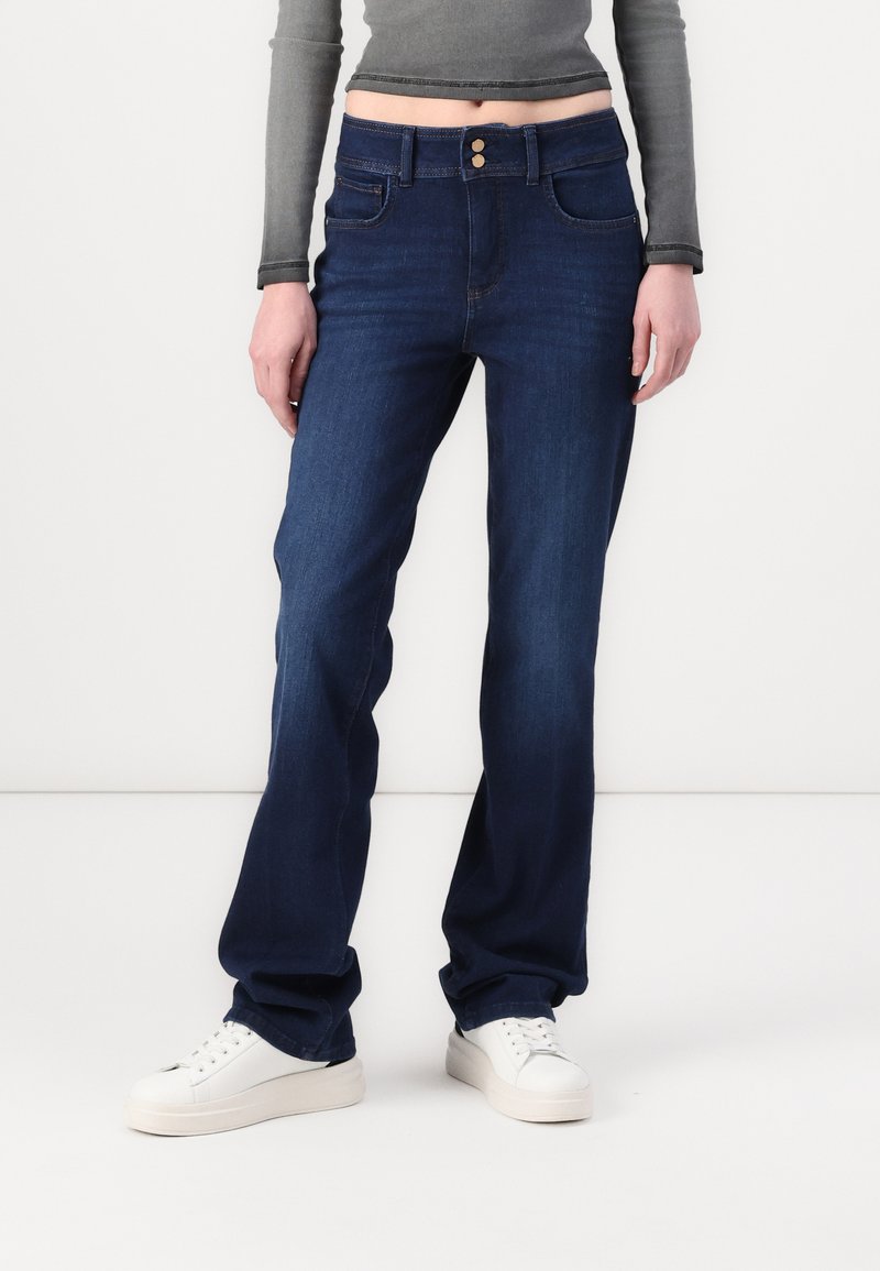 Blue denim jeans with a classic cut and two buttons at the waist. Features front pockets and a smooth texture, paired with white sneakers.