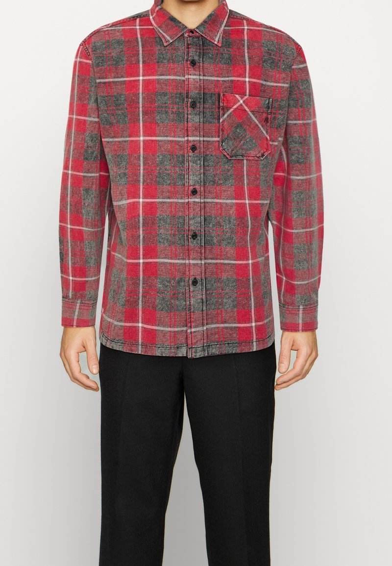Homme portant une chemise à manches longues à carreaux rouge, gris et blanc avec une poche poitrine et un pantalon noir.