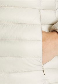 Światło szara tkanina kurtki ocieplanej o gładkiej fakturze, z poziomymi przeszyciami i boczną kieszenią na zamek.