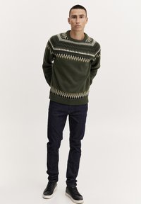 Grüner Strickpullover mit geometrischem Muster in Schwarz, Weiß und Creme am Hals und Saum; kombiniert mit dunklen Denim-Jeans und schwarzen Sneakern.