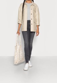 Persona vestida con chaqueta beige, camisa gris, jeans ajustados negros, zapatillas blancas, sosteniendo un bolso de mano beige y blanco con estampado, de pie sobre un fondo sencillo.