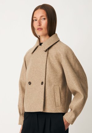 Femme portant une veste en laine beige à double boutonnage avec des manches larges et un pantalon noir sur un fond uni.