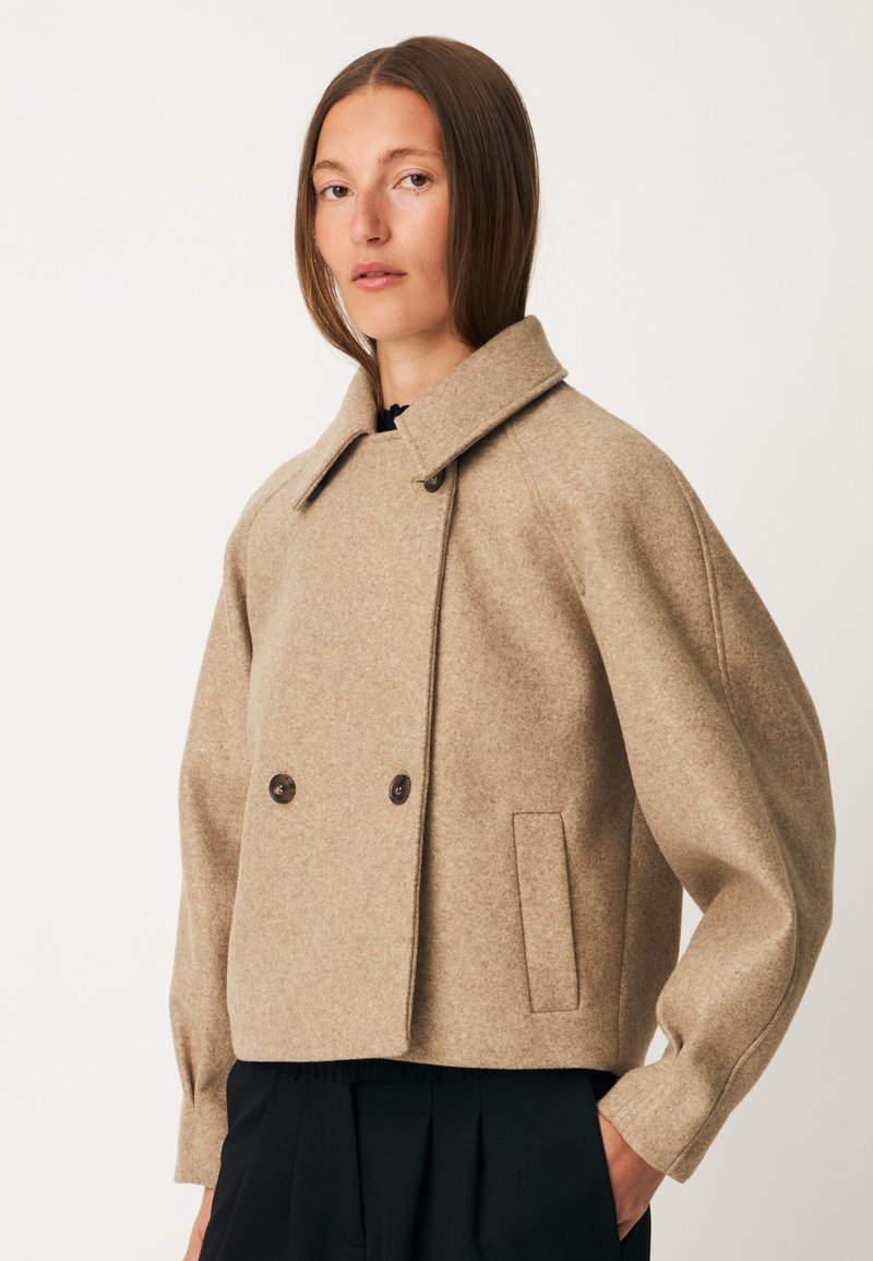 Femme portant une veste en laine beige à double boutonnage avec des manches larges et un pantalon noir sur un fond uni.